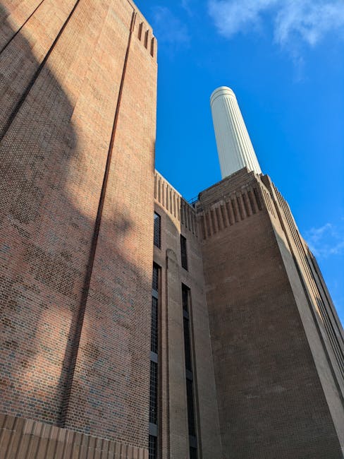 A view of the Battersea Power Station area featuring the industrial-style building with four tall chimneys, constructed from red brick, situated near the river with boats docked along the shoreline. A