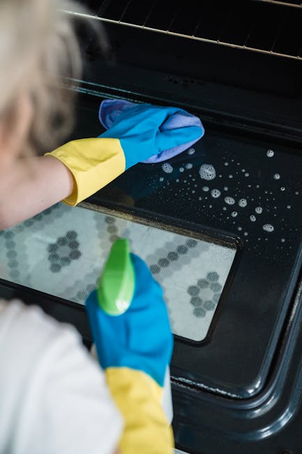 A person wearing blue and yellow rubber cleaning gloves is cleaning the glass window of an oven. The oven's interior, visible through the glass, appears to be dark with some soap suds and bubbles indi
