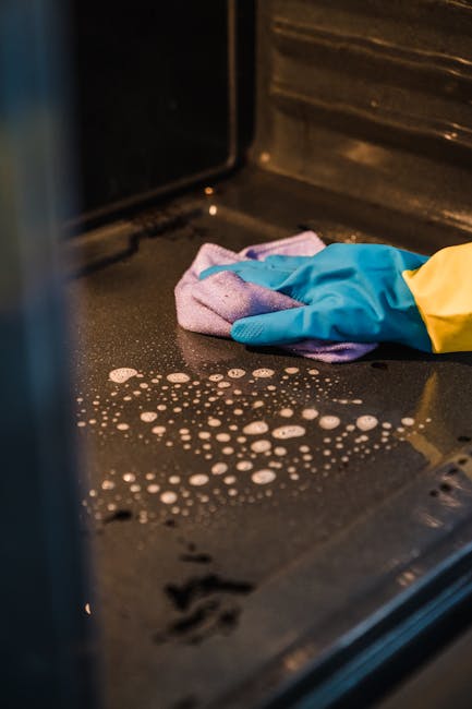 A person wearing blue and yellow rubber cleaning gloves is cleaning the glass window of an oven. The oven's interior, visible through the glass, appears to be dark with some soap suds and bubbles indi