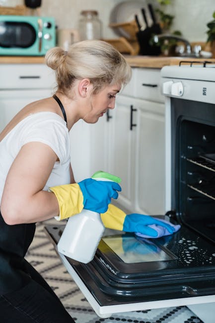 Close-up of a person wearing yellow and blue cleaning gloves, adjusting a wire oven rack inside a black oven. The oven's interior, featuring a clean, stainless steel surface, is visible with a well-li